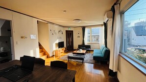 Un soggiorno moderno con pavimento in legno, un divano, un tavolino da caffè e una televisione. Si vede una scala che conduce a un altro livello, e una finestra con vista sugli edifici circostanti.