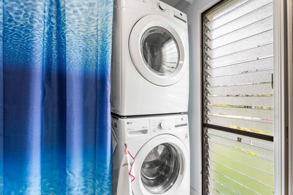 A laundry area with two stacked washing machines, a blue curtain with a water pattern, and a window with blinds.