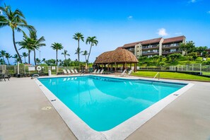 Una piscina con sillones de descanso, un pabellón de techo de paja y un hotel al fondo.