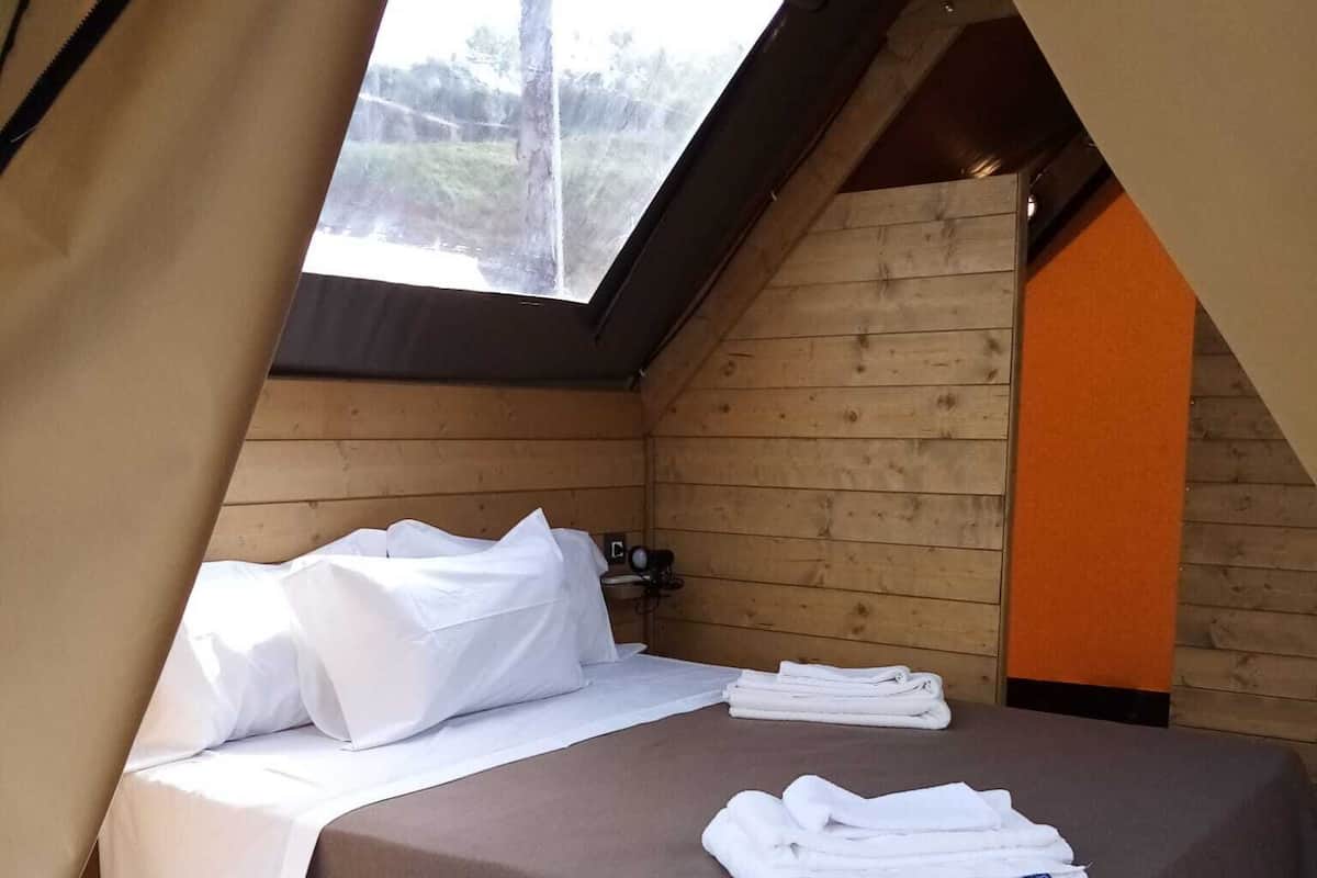 A single bed with white linens and a brown bedspread in a tent-style room with wooden walls and a skylight.