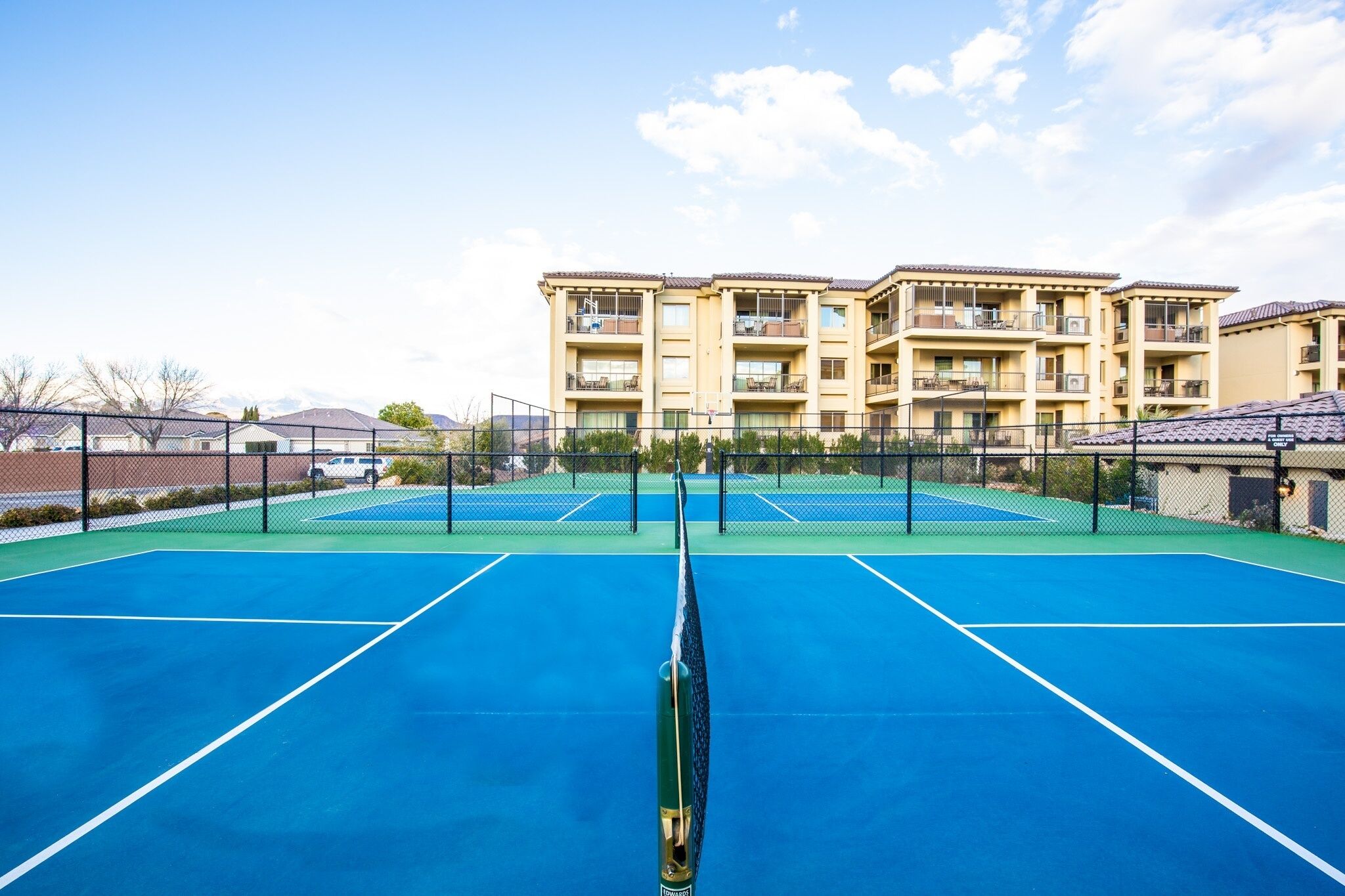 A tennis court with a net in the center, surrounded by a fence. In the background, there is a multi-story building with balconies.
