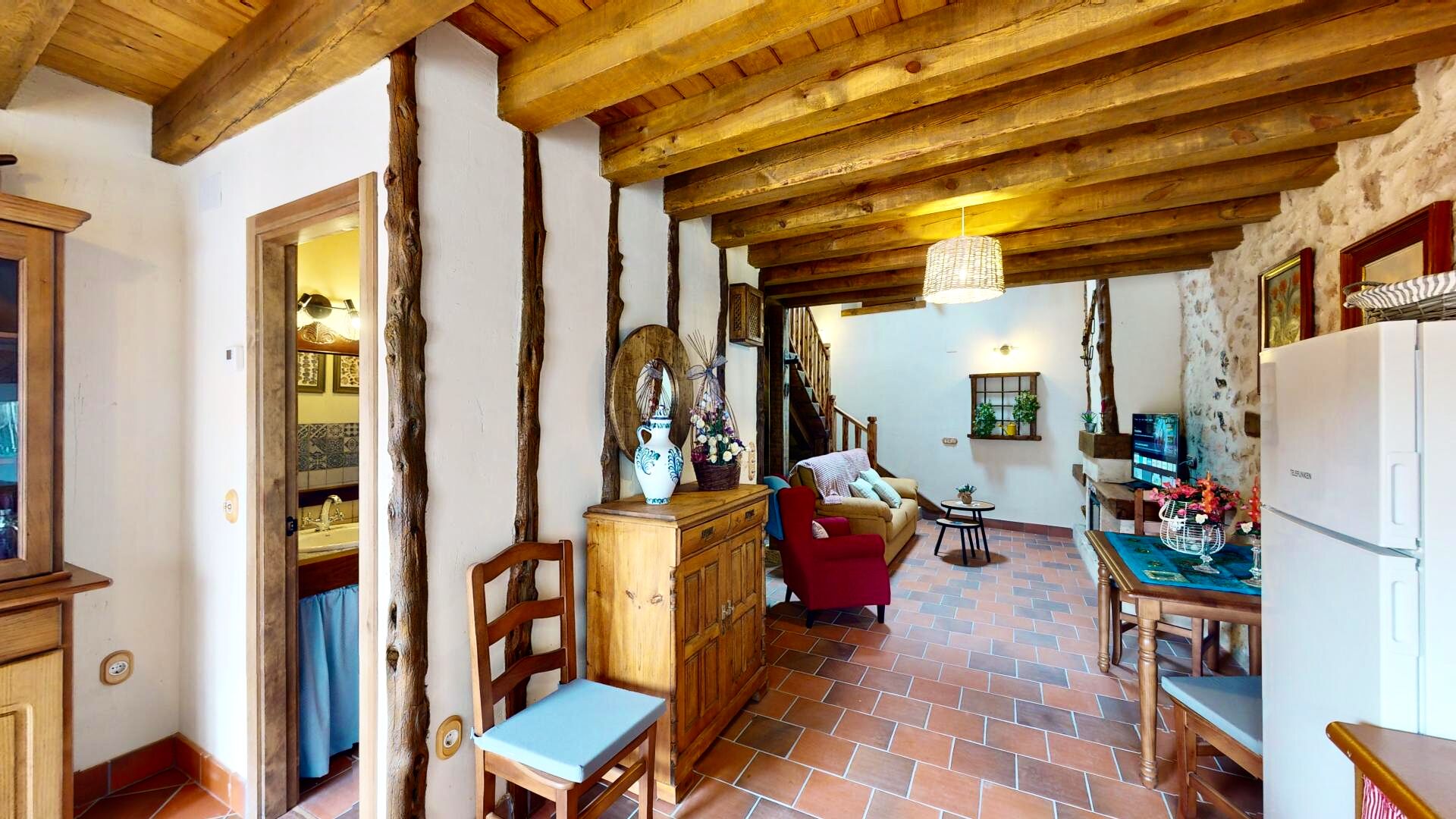 A rustic interior with exposed wooden beams, a tiled floor, a wooden dining table, and a red armchair.