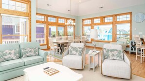 A spacious living room with a light blue sofa, two armchairs, a coffee table, and a dining area with wooden chairs and a chandelier.