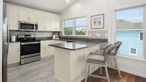 A modern kitchen with white cabinets, stainless steel appliances, a granite countertop, and a large window with a view of a blue house and hills. - Luxury Home Near Beach with Pool, Spa! (Corolla)