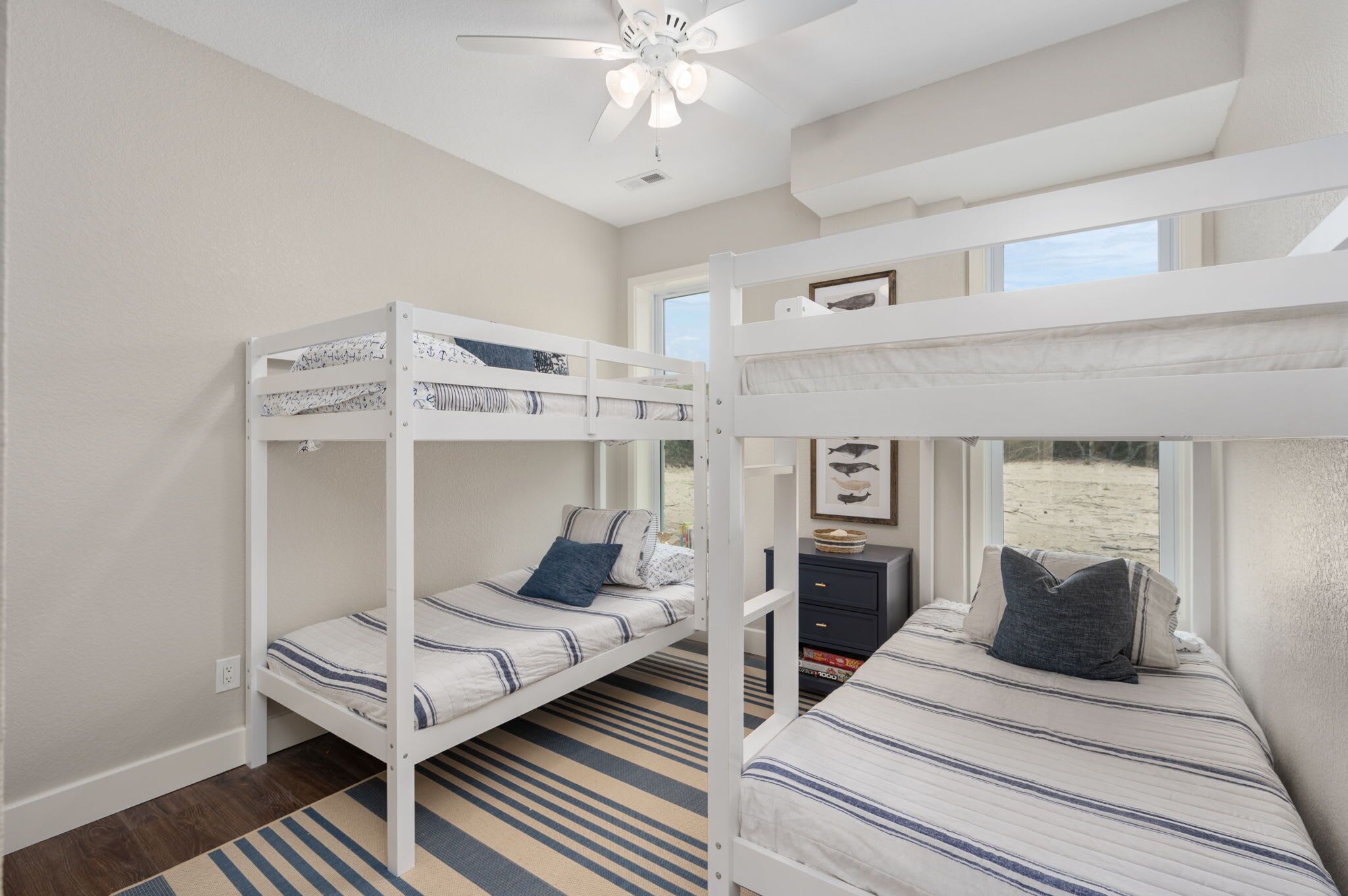 A room with two bunk beds, a ceiling fan, a window with a beach view, and a small desk with a lamp.