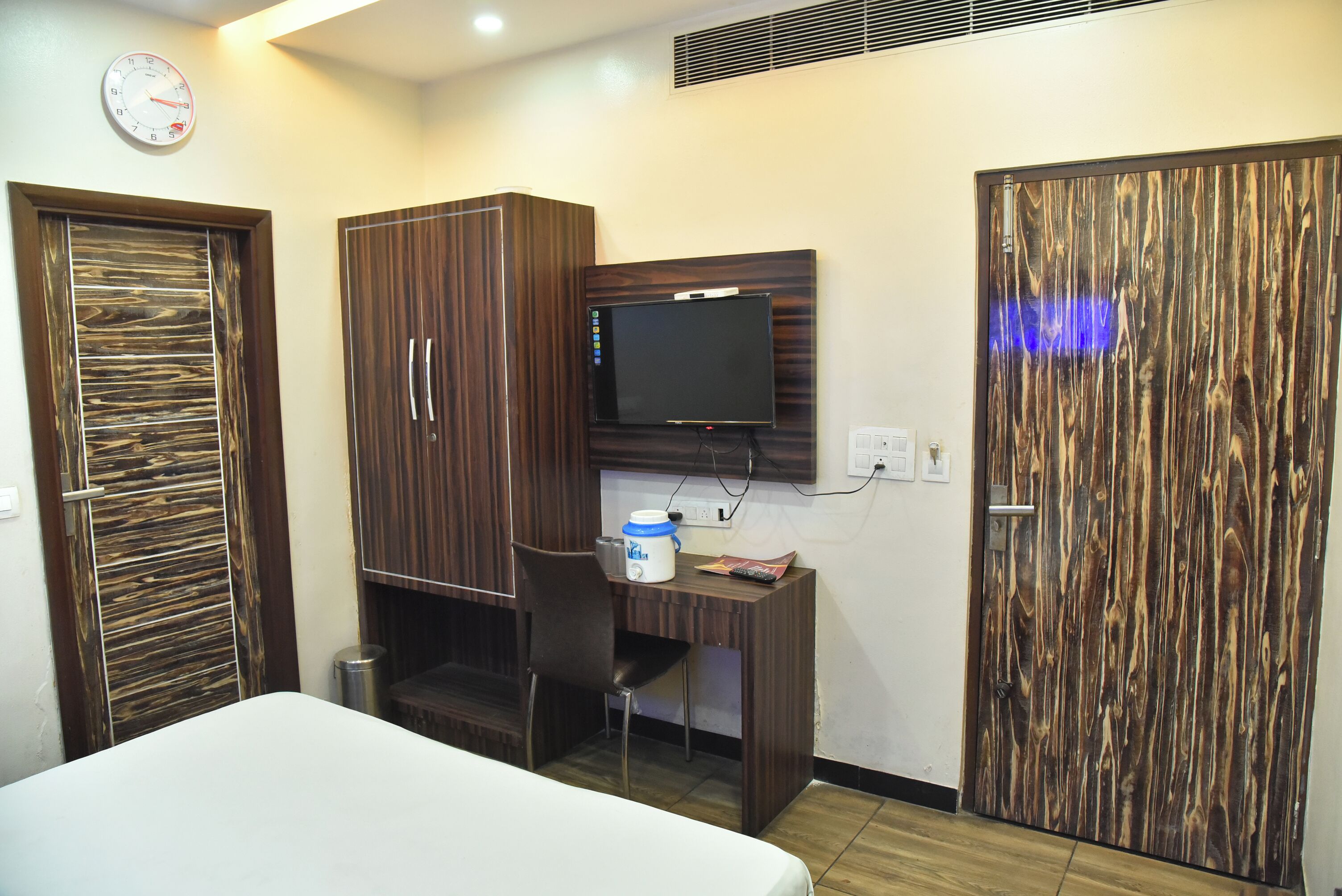 A hotel room with a bed, a desk with a computer, a TV, and two wooden doors.