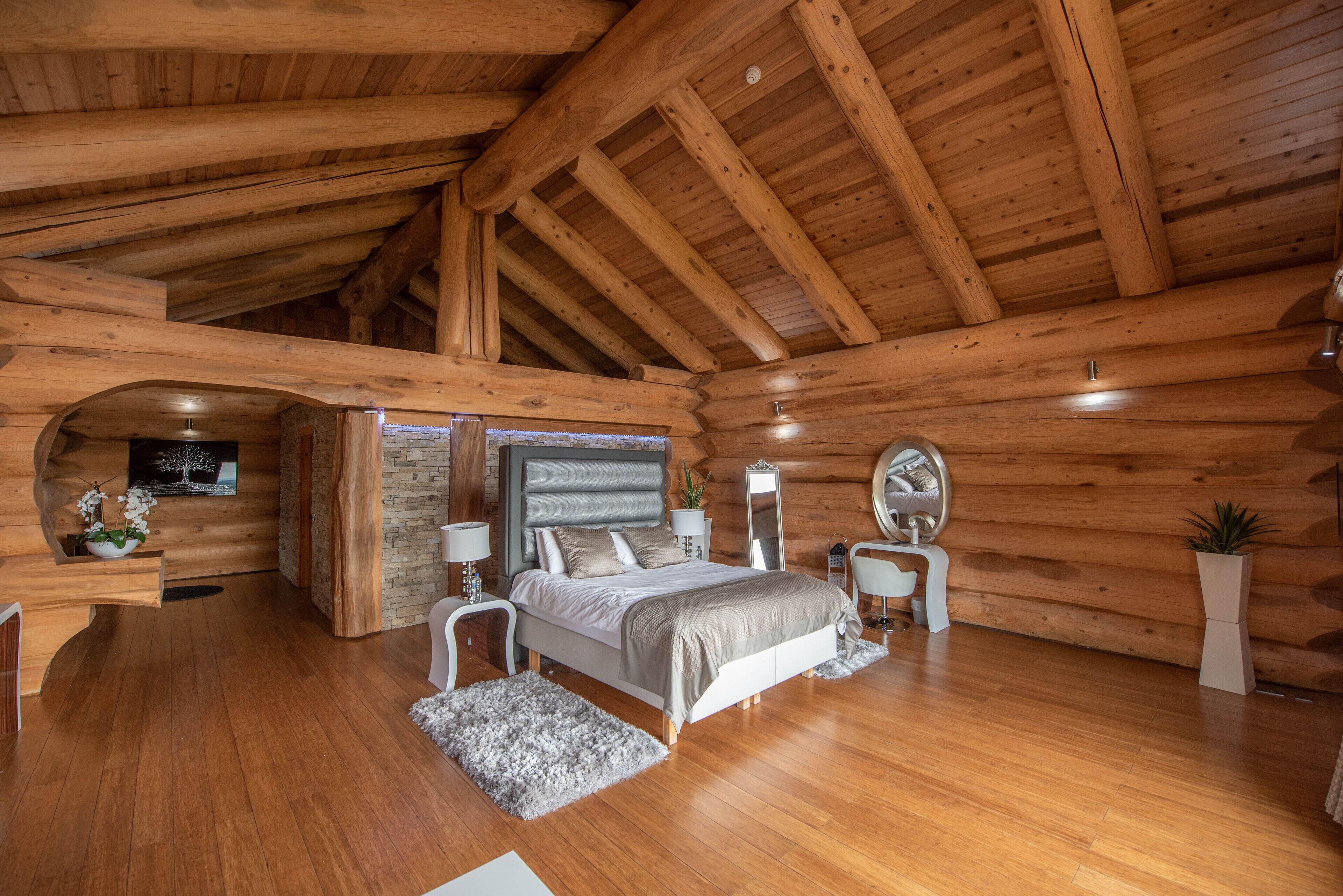 A log cabin bedroom with a wooden ceiling, a bed with a headboard, bedside tables, a mirror, and a small desk with a chair.