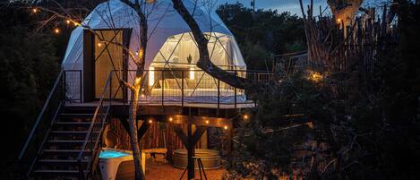 A dome-shaped cabin with a staircase leading to a deck, surrounded by trees and a fire pit.