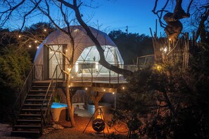 A dome-shaped cabin with a staircase leading to a deck, surrounded by trees and illuminated by string lights.