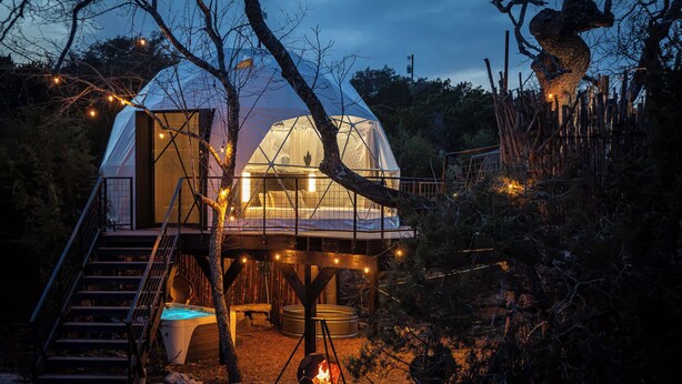 A dome-shaped cabin with a staircase leading to a deck, surrounded by trees and a fire pit.
