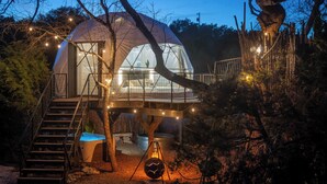 A dome-shaped cabin with a staircase leading to a deck, surrounded by trees and illuminated by string lights.