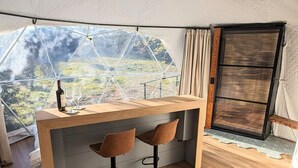 A compact kitchen area with a wooden countertop, two bar stools, and a large window offering a view of a green landscape. - Treehouse!! Serenity Dome With Private Hot Tub! (Fredericksburg)