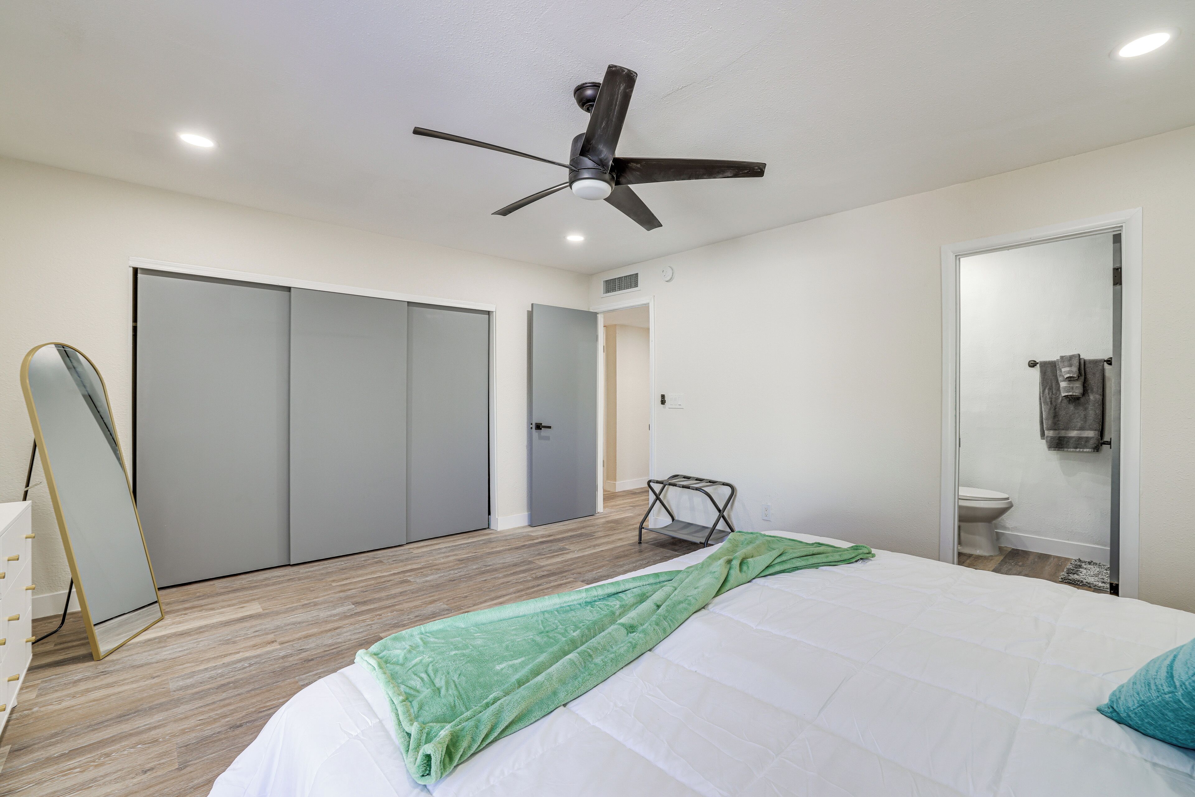 Un dormitorio con cama, ventilador de techo, armario, espejo y baño con inodoro y lavabo.