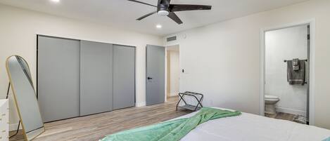 Un dormitorio con cama, ventilador de techo, armario, espejo y baño con inodoro y lavabo.