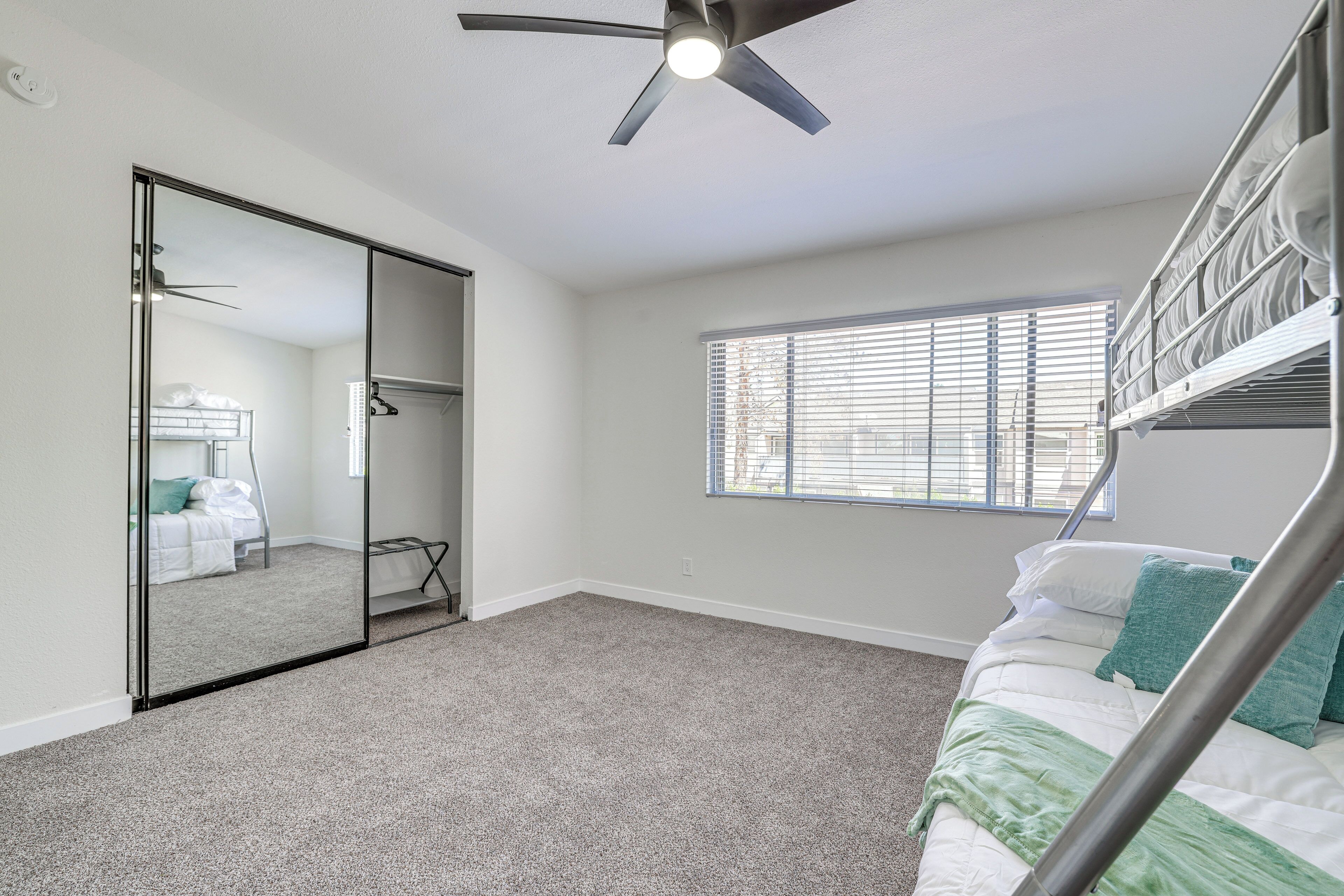 A bedroom with a bunk bed, a ceiling fan, a large mirror, and a window with blinds.