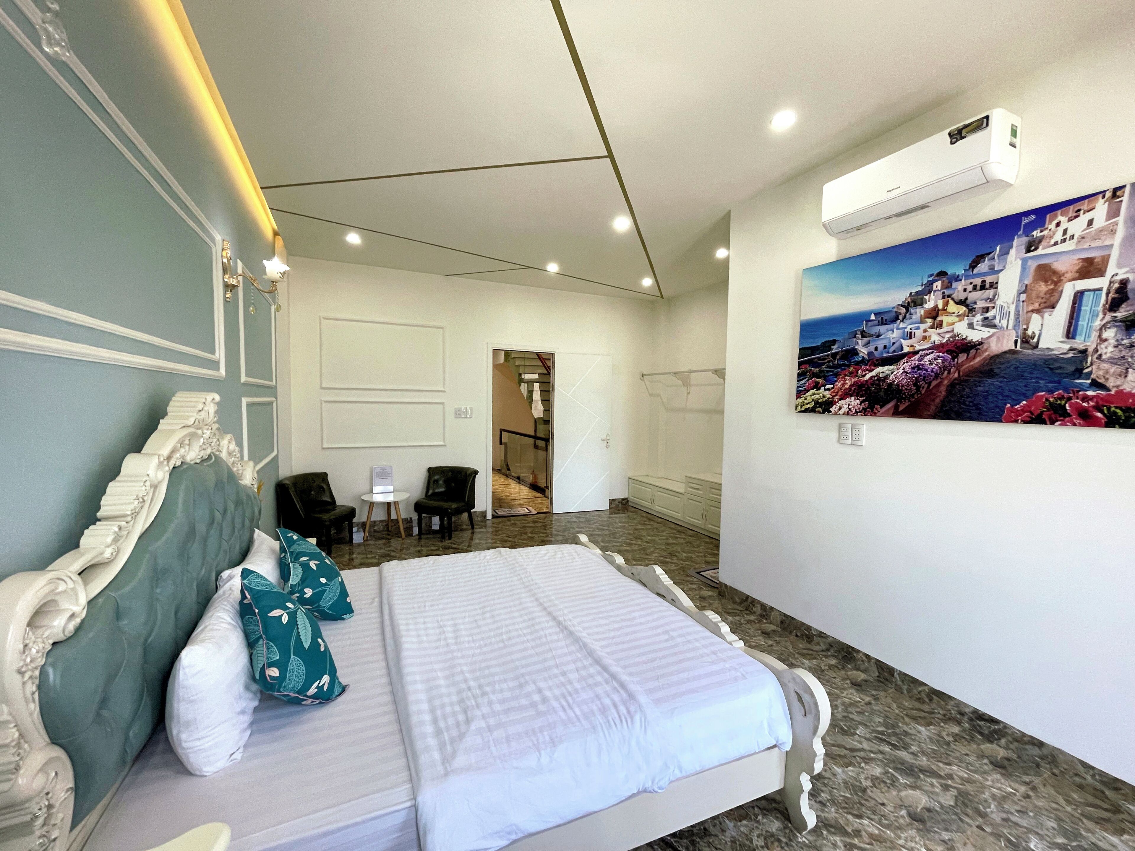 Una habitación de hotel con una cama grande, una mesita pequeña, dos sillas, un cuadro de una escena costera y un equipo de aire acondicionado.