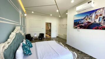 Una habitación de hotel con una cama grande, una mesita pequeña, dos sillas, un cuadro de una escena costera y un equipo de aire acondicionado.