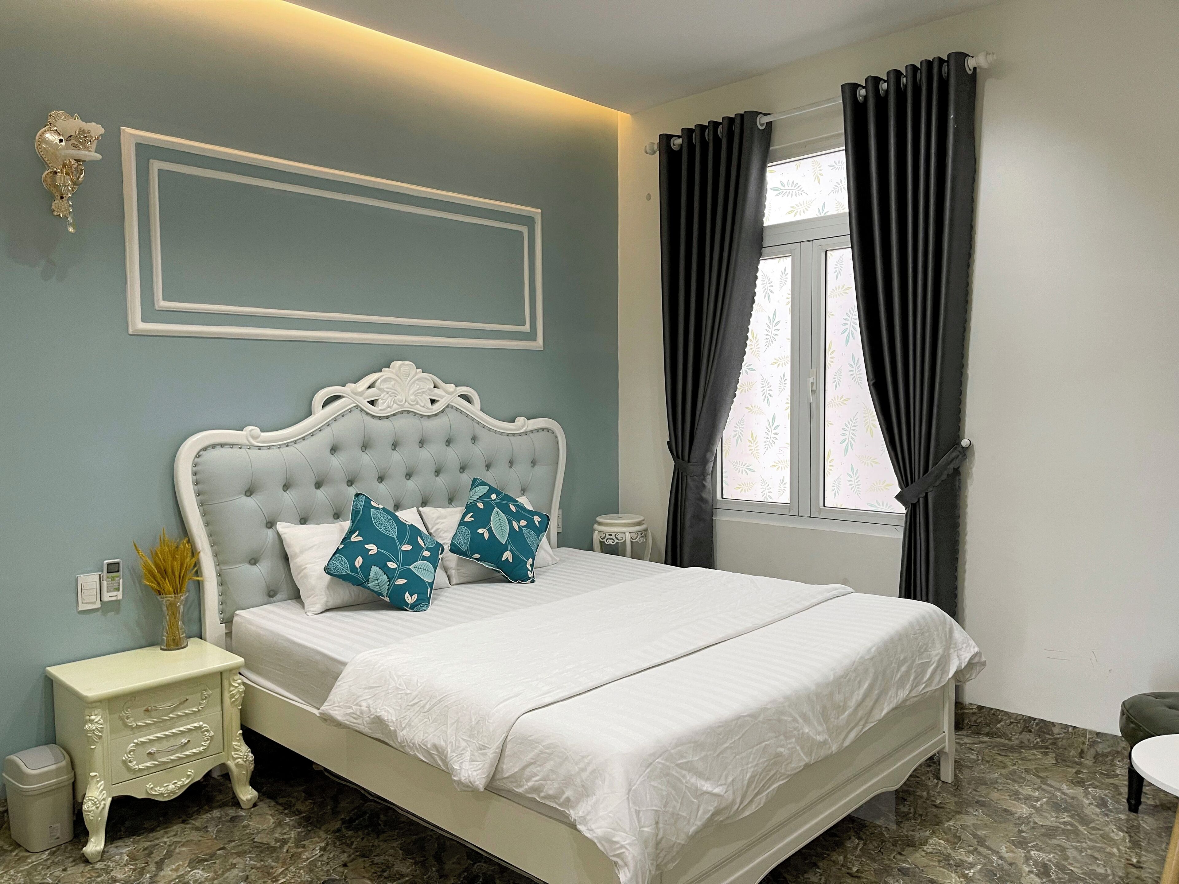 A neatly arranged bedroom with a tufted headboard, a bedside table with drawers, a small stool, and a window with curtains.