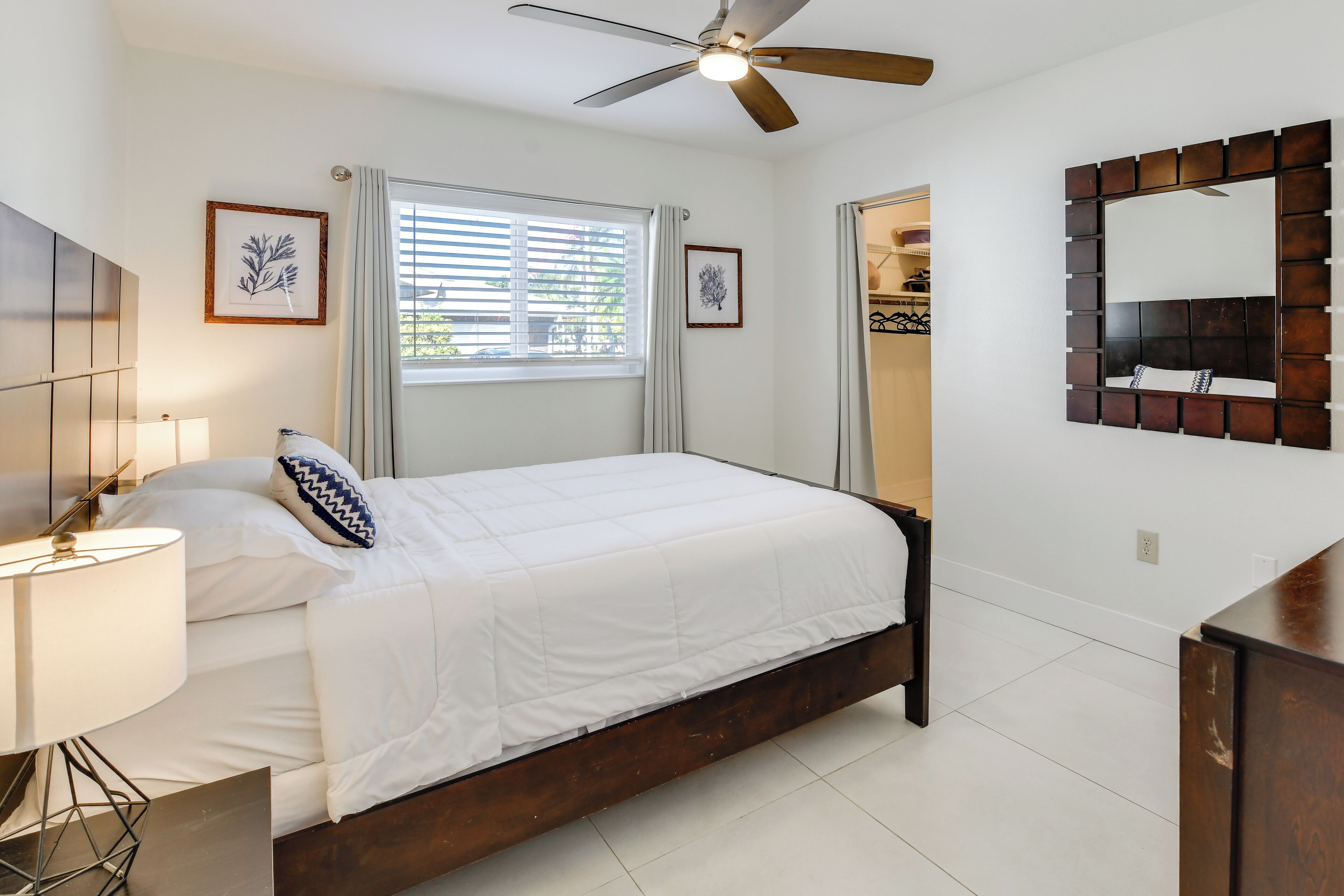 A bedroom with a bed, a ceiling fan, a window with blinds, a bedside lamp, and a mirror on the wall.