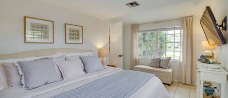 Ein Schlafzimmer mit einem großen Bett, einem Nachttisch mit Lampe, einem Fenster mit Vorhängen und zwei gerahmten Bildern an der Wand.