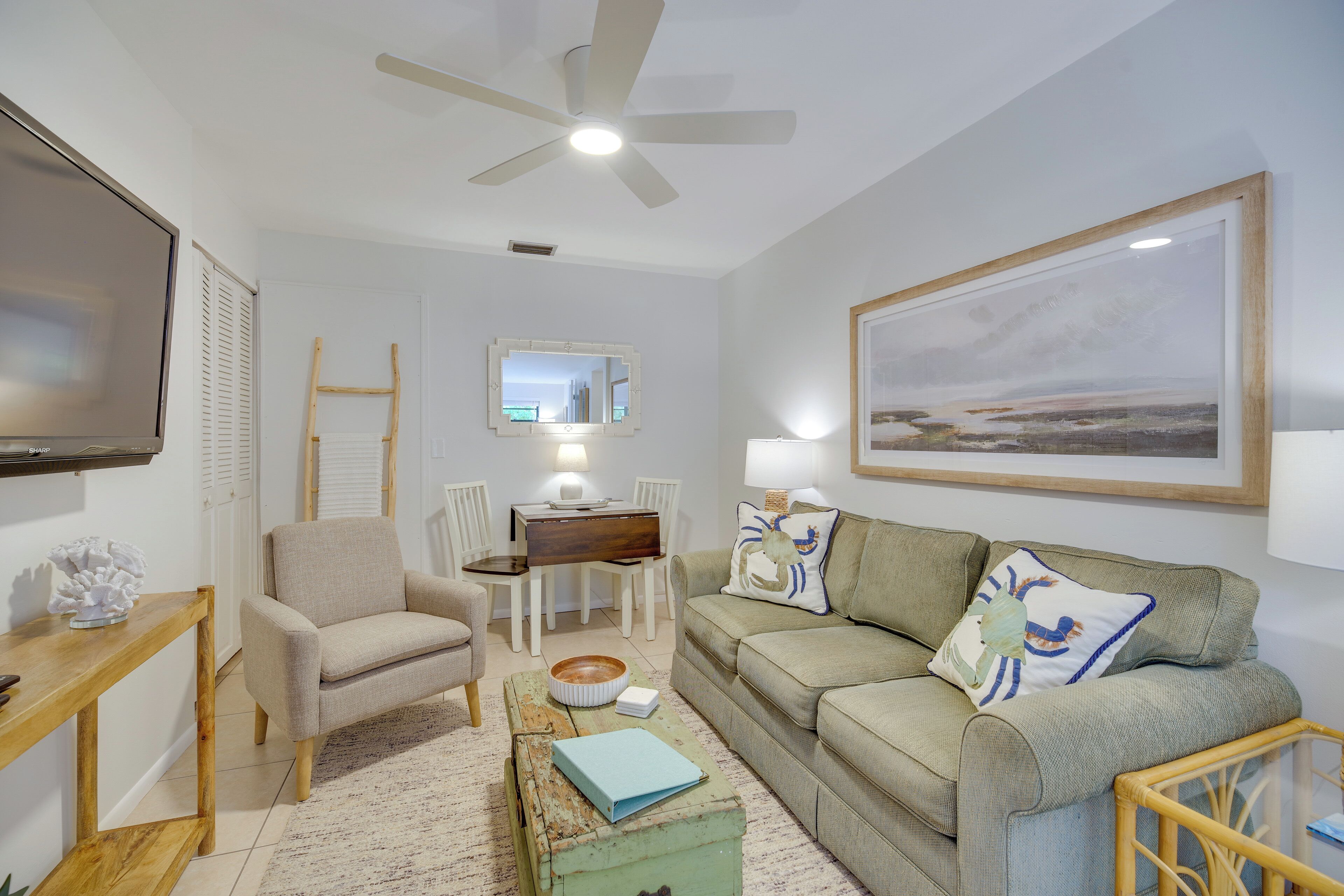 A living room with a sofa, armchair, coffee table, and a TV. There is a dining table with chairs and a painting on the wall.