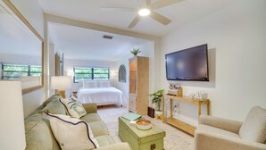 A well-lit living room with a large flat-screen TV mounted on the wall, a comfortable sofa, a coffee table, and a bed in the background. - Vero Beach Studio w/ Shared Patio ~ 3 Mi to Beach! (Vero Beach)