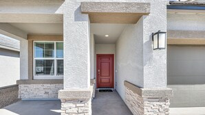 A building entrance with a red door, stone pillars, and a concrete pathway leading to a garage. - Maricopa Home w/ Putting Green & Covered Patio! (Maricopa)