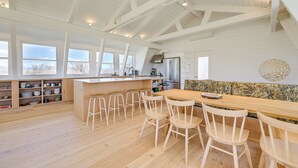 Een ruime keuken met een houten bar en krukken, grote ramen en een hoog plafond met zichtbare balken.