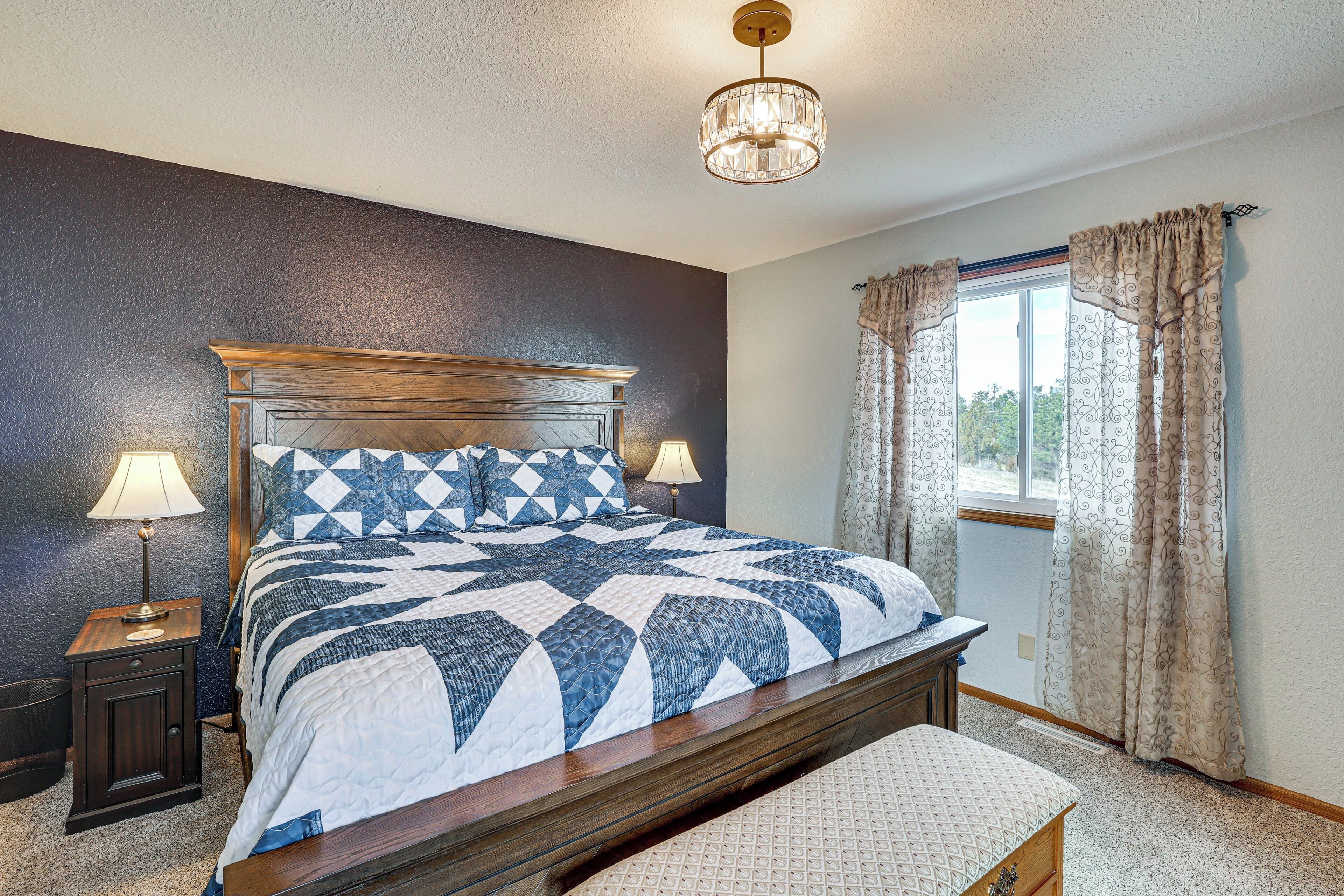 A bedroom with a large bed, a nightstand, a bedside lamp, a window with curtains, and a chandelier.