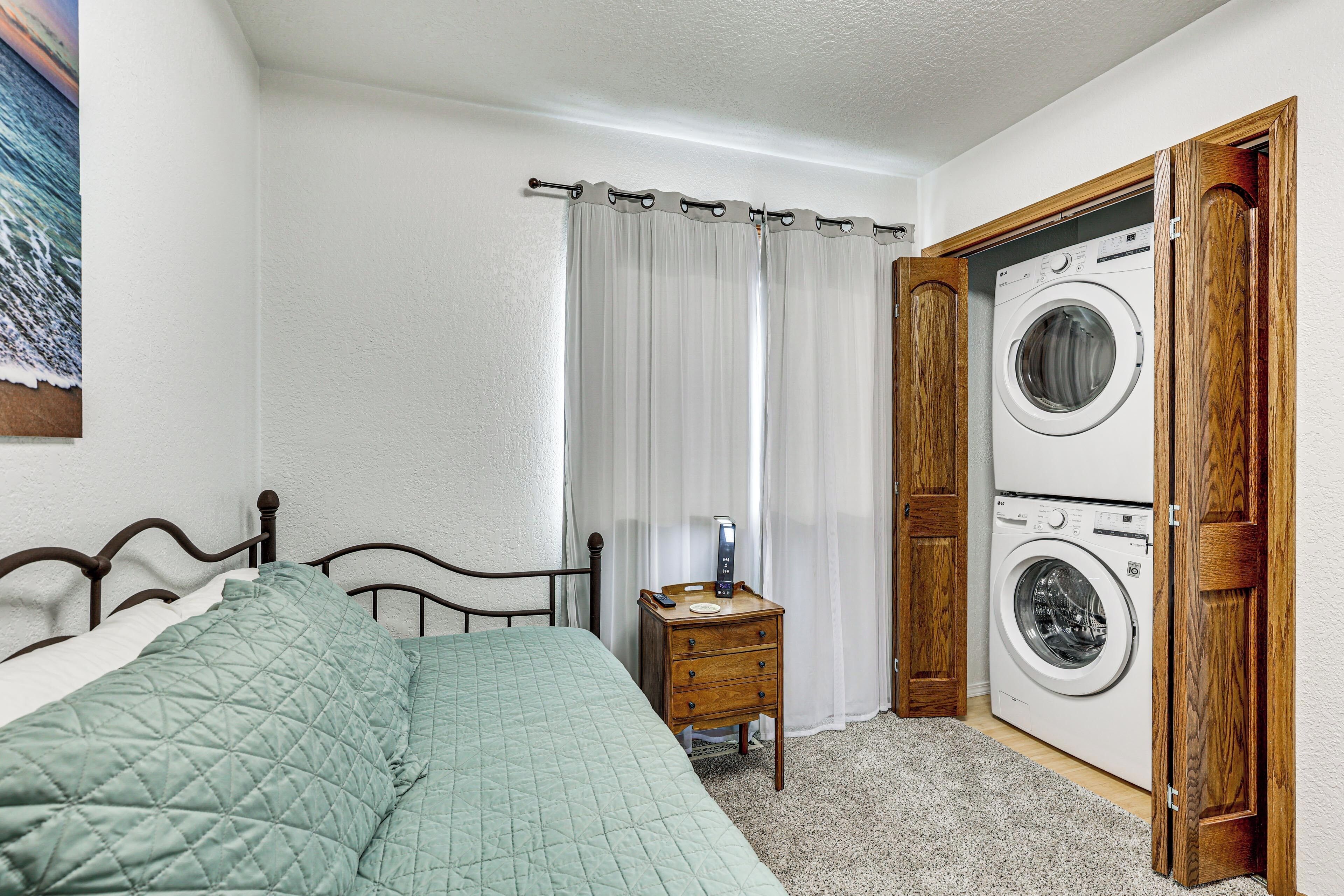Un dormitorio con una cama, una mesita de noche de madera con un teléfono y un electrodoméstico, y una lavadora dentro de un mueble de madera.