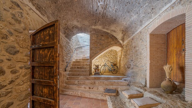 A stone-walled interior with a wooden door, a staircase, and a bicycle leaning against the wall.