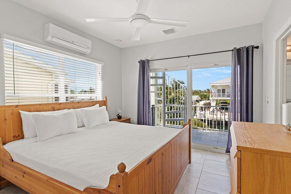 A bedroom with a wooden bed, a ceiling fan, an air conditioning unit, a balcony with a view of the surroundings, and a wooden dresser.