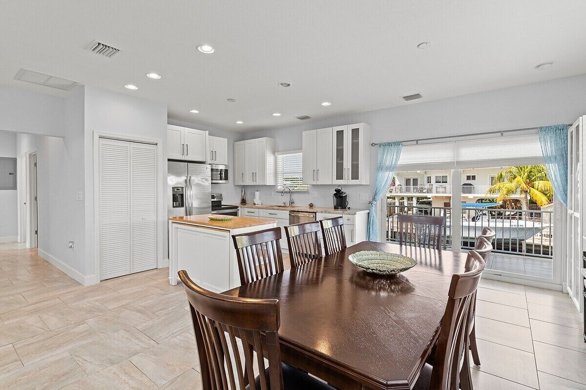 A modern kitchen with white cabinets, a wooden dining table, and a balcony with a view of palm trees.