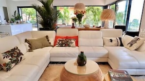 Uma sala de estar moderna com um sofá seccional branco, uma mesa de centro redonda trançada e vista para o exterior através de amplas janelas.