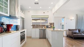 Een moderne keuken met witte kasten, een ingebouwde oven en een vaatwasser. Er is een klein eiland met een magnetron en een spoelbak. In de woonkamer is een bruine leren bank zichtbaar.