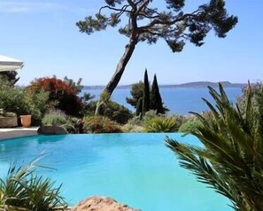 A pool with clear blue water, surrounded by various plants and trees, with a view of the sea and distant land in the background. - Magnifique Villa, Piscine à Débordement, vue mer (Hyères)