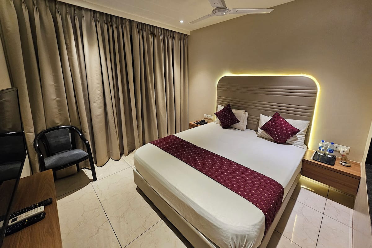 Habitación de hotel con una cama grande, ventilador de techo, televisor, una silla y una mesita de noche con lámpara y botellas de agua.