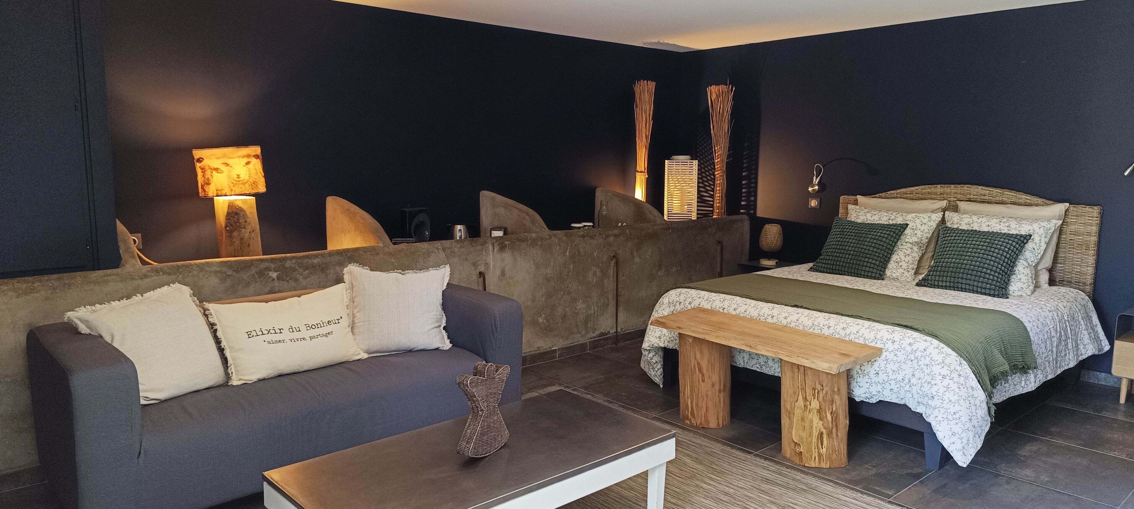 A modern bedroom with a dark blue wall, a grey sofa with decorative pillows, a wooden bed frame, and a bedside table.