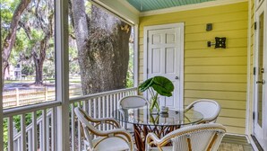 A covered porch with wicker chairs and a glass table, a white door, and a large tree outside. - Two-Bedroom, Two-Bath, Single-Story Cottage Located in Wachesaw Plantation (Murrells Inlet)