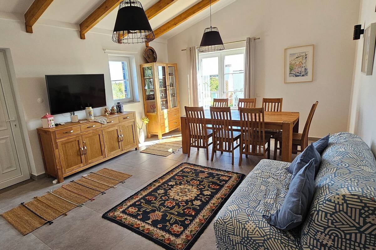 A spacious living room with a sofa, a dining table with chairs, a TV, wooden cabinets, and a large window.