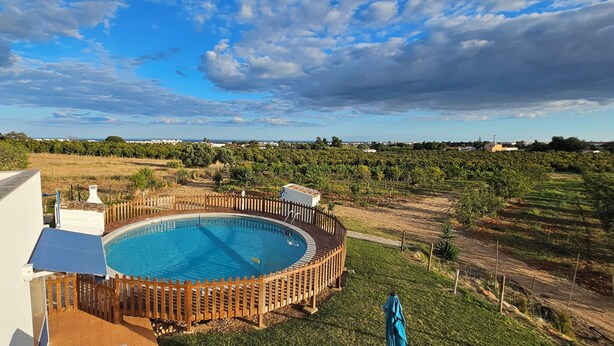 A circular pool with a wooden fence, surrounded by a grassy area and trees, with a view of distant buildings and a cloudy sky. - Quinta da Bornacha (Vila Real Santo Antonio)
