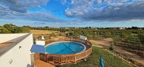 A circular pool with a wooden fence, surrounded by a grassy area and trees, with a view of distant buildings and a cloudy sky. - Quinta da Bornacha (Vila Real Santo Antonio)