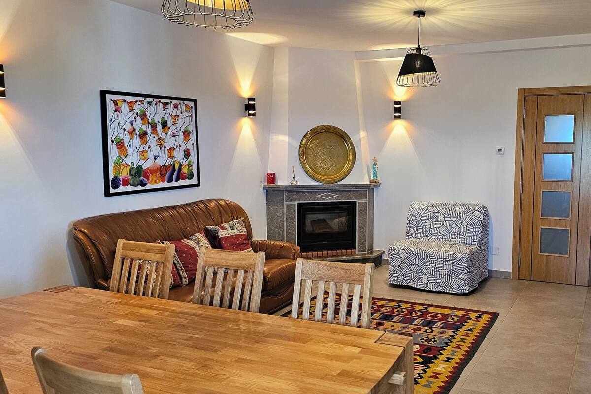 A cozy living room with a brown leather sofa, a patterned armchair, a wooden dining table, and a fireplace.