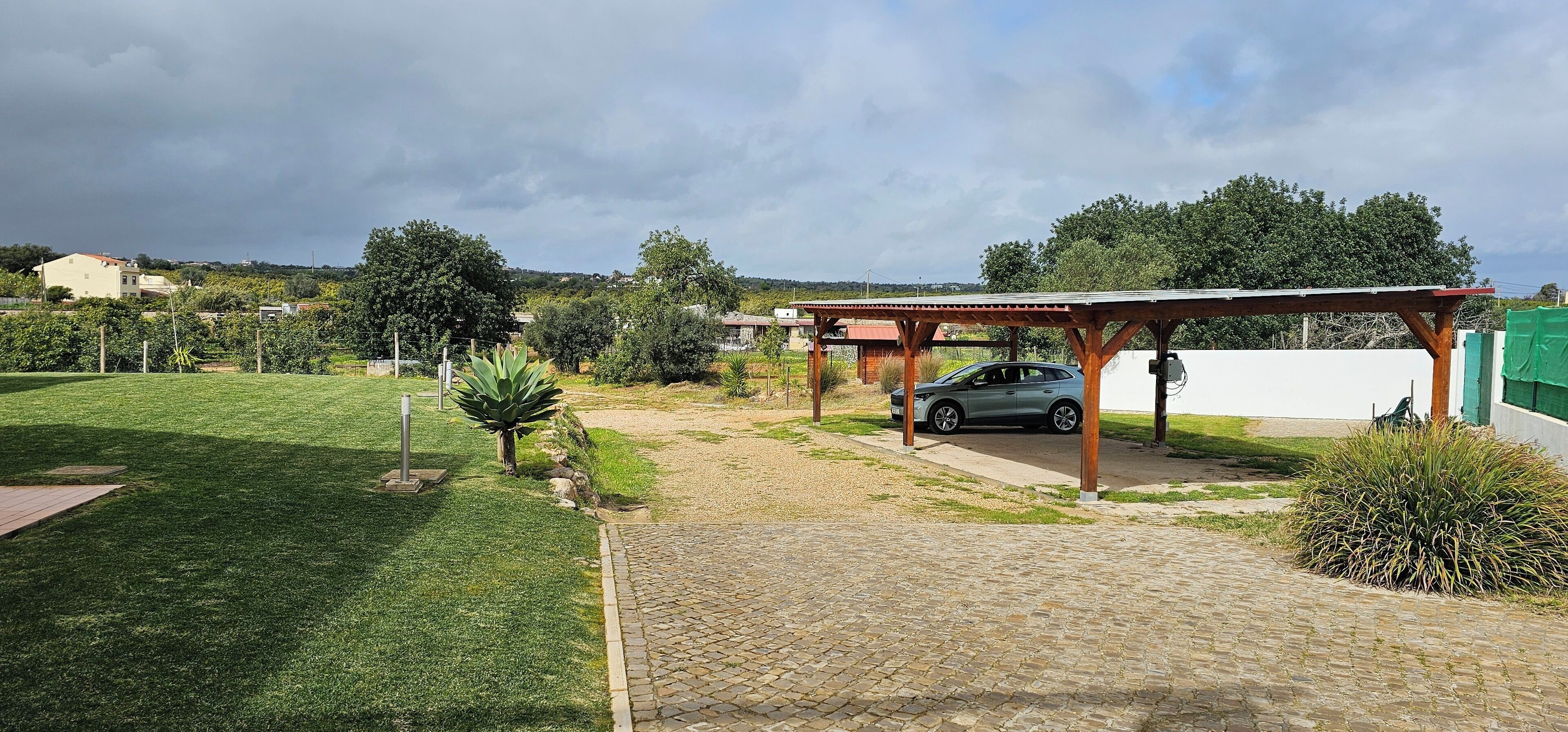 A paved driveway leading to a covered car parking area with a car parked under a wooden pergola.