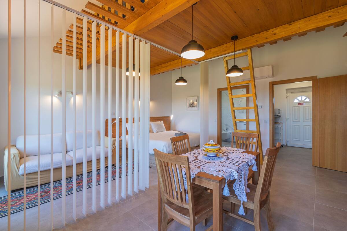 A cozy room with a wooden ceiling, a dining table with chairs, a bed with a white headboard, and a white partition with vertical slats.