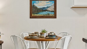 A dining table with ceramic dishes and a potted plant, surrounded by white chairs, with a framed painting above.