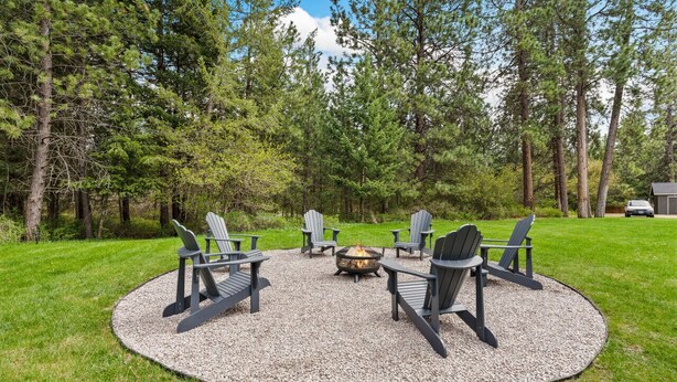 A circular outdoor seating area with Adirondack chairs around a fire pit, surrounded by a gravel circle on a grassy lawn, with trees in the background.