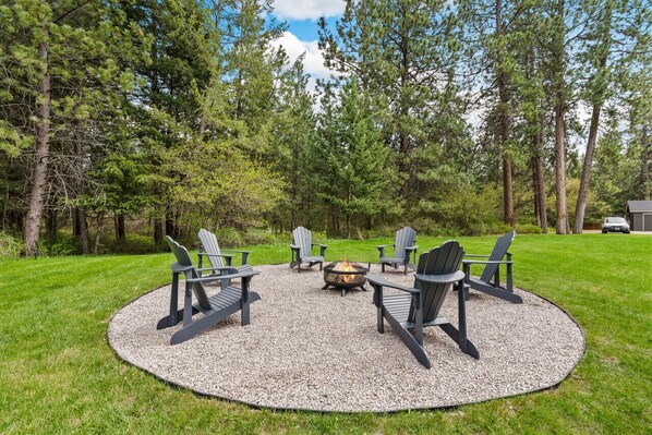 Un espace extérieur circulaire aménagé avec des chaises Adirondack disposées autour d’un foyer, le tout entouré d’un cercle de gravier sur une pelouse verdoyante, avec des arbres en arrière-plan.