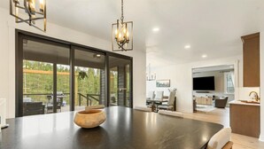 A modern kitchen with a large island, a dining area with a table and chairs, a TV mounted on the wall, and a view of the outdoors through large sliding doors. - Quiet Neighborhood Home w/ Pristine Mountain Views (Kalispell)