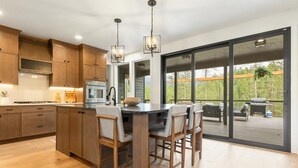 A modern kitchen with wooden cabinets, a central island with bar stools, and a sliding glass door leading to an outdoor patio. - Quiet Neighborhood Home w/ Pristine Mountain Views (Kalispell)